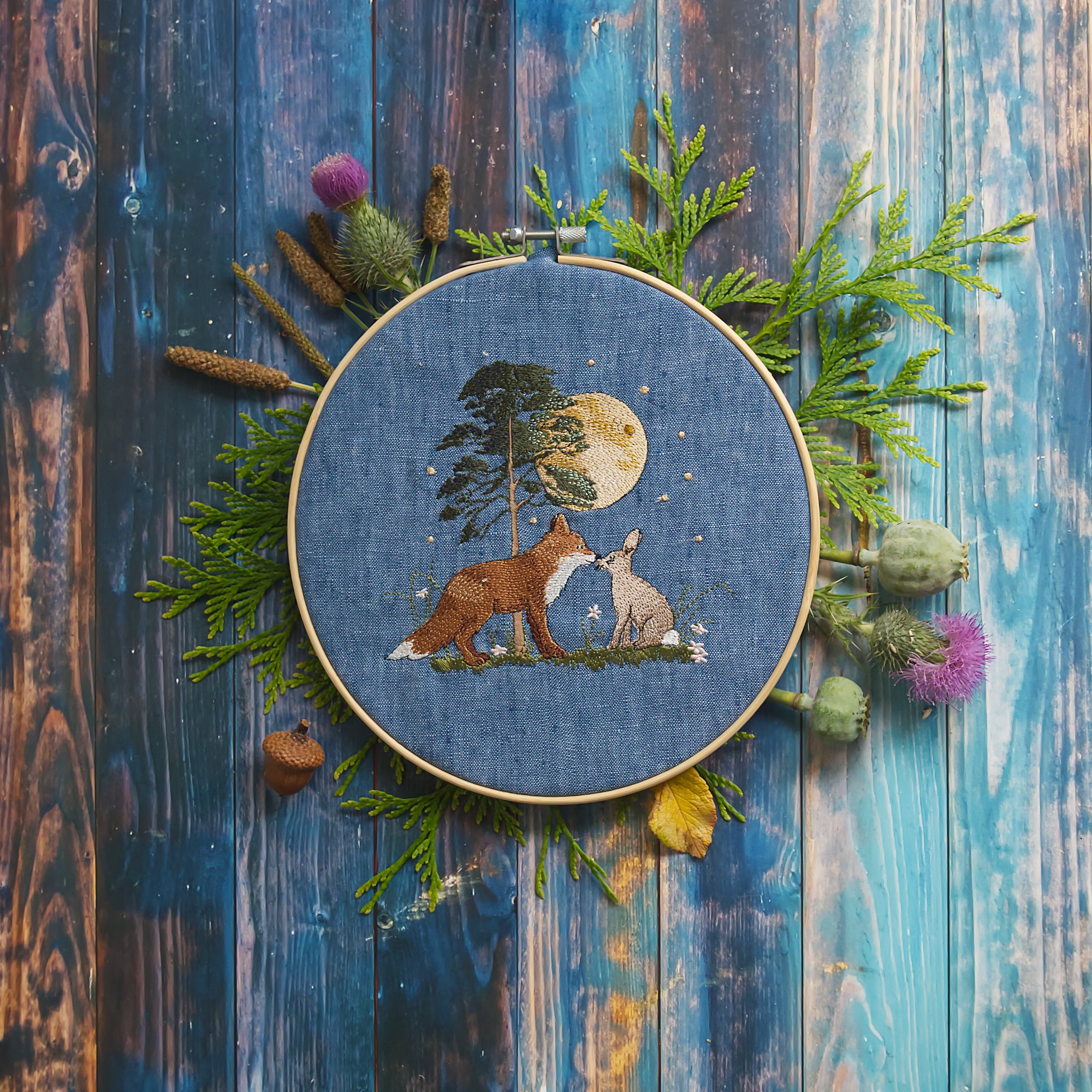Stickdatei  Szene mit Fuchs und Hase unter einem leuchtenden Vollmond vor einem Baum, liebevoll inszeniert im Stickrahmen auf blauem Stoff und dekoriert mit Naturmaterialien wie Farn, Disteln und Mohnkapseln.