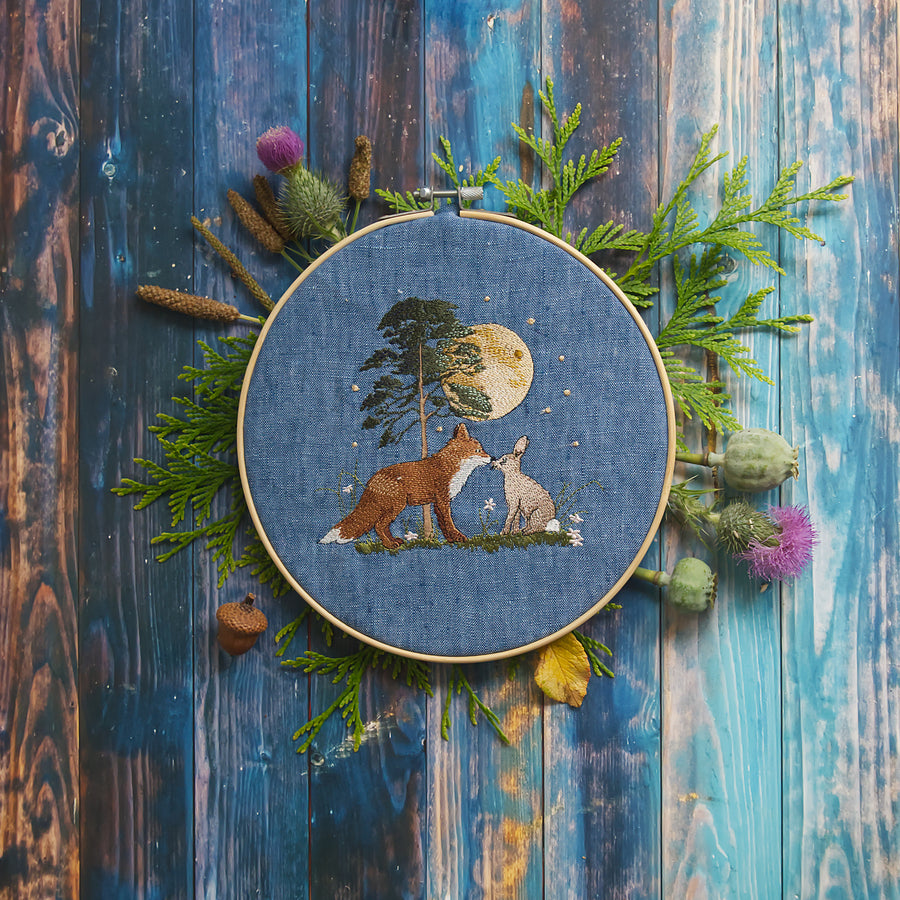 Stickdatei  Szene mit Fuchs und Hase unter einem leuchtenden Vollmond vor einem Baum, liebevoll inszeniert im Stickrahmen auf blauem Stoff und dekoriert mit Naturmaterialien wie Farn, Disteln und Mohnkapseln.