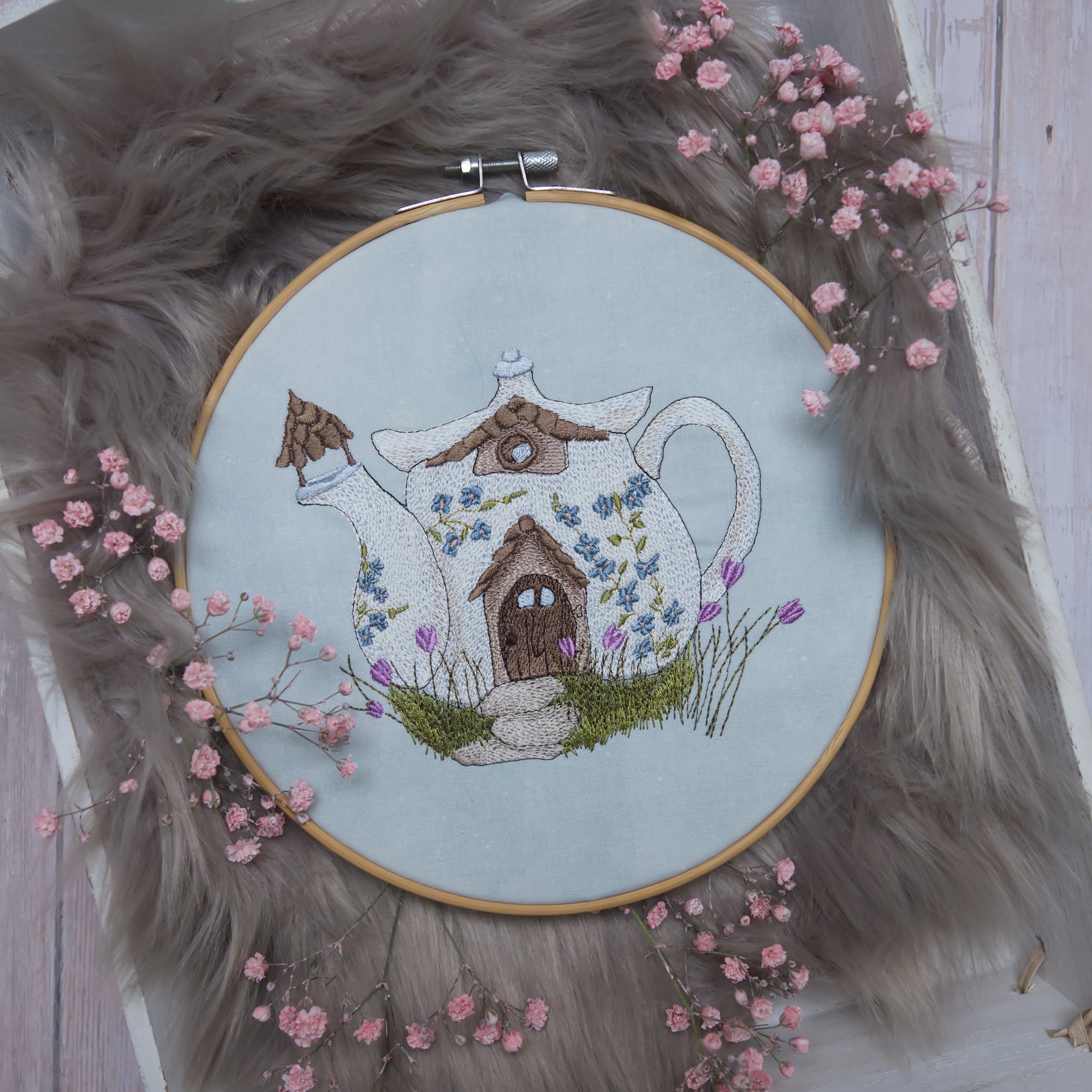 Detailreiche Stickerei eines märchenhaften Teekannenhauses mit Blumen auf hellem Stoff im Stickrahmen, frühlingshafte Landhaus-Dekoration.