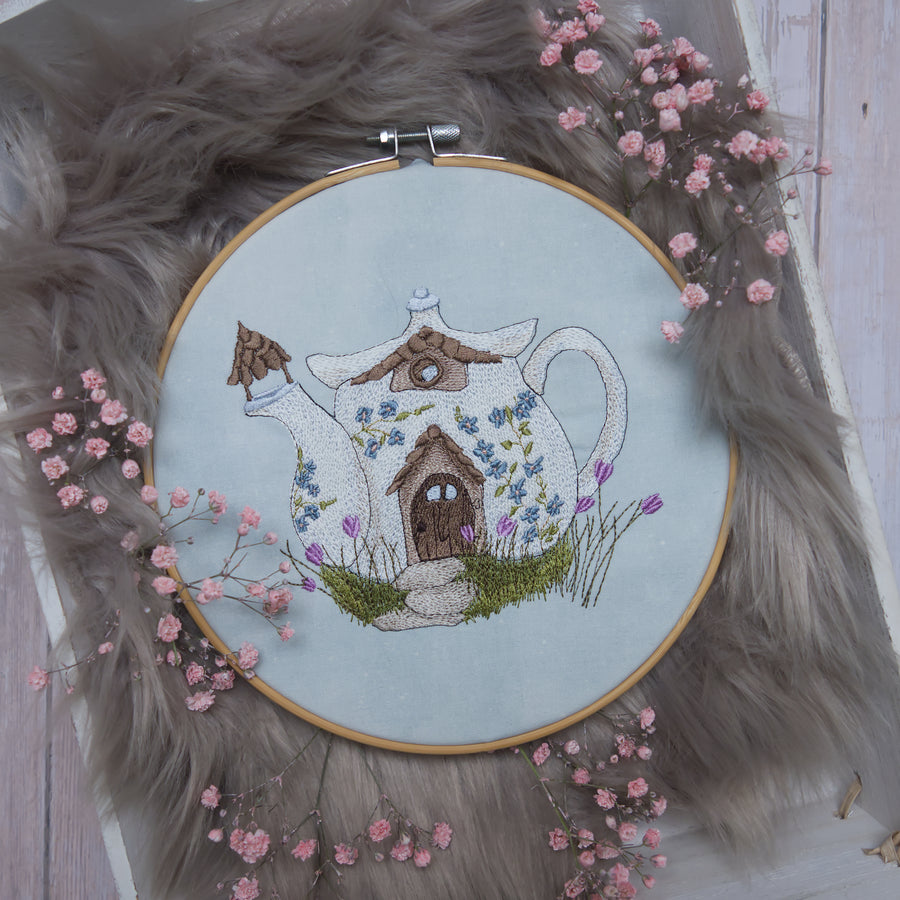 Detailreiche Stickerei eines märchenhaften Teekannenhauses mit Blumen auf hellem Stoff im Stickrahmen, frühlingshafte Landhaus-Dekoration.